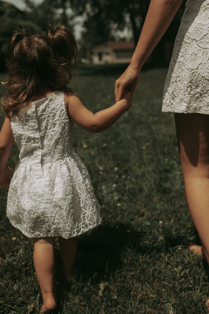 Una niña pequeña con vestido blanco toma la mano de su mamá, de espaldas (foto de demostración)