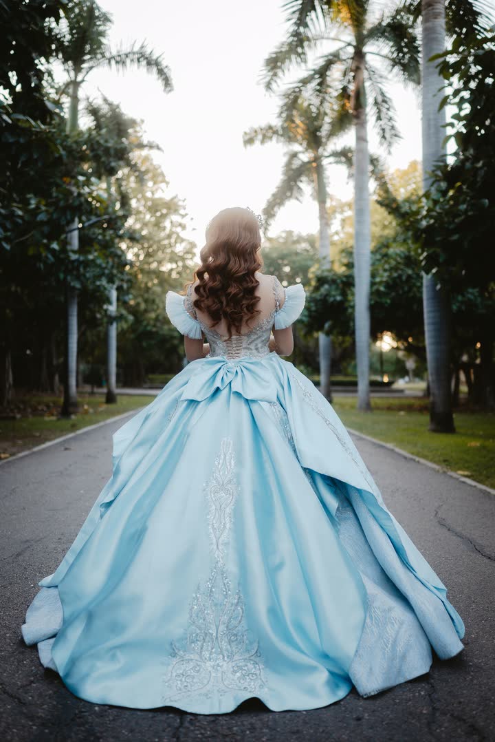 La quinceañera de espaldas con un vestido azul claro, en un camino de palmeras (foto de demostración)